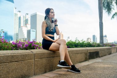 Asyalı kadın bahçedeki egzersizden sonra dinlenmek için çiçeğin yanına oturur ve arka planda büyük bir şehir inşa eder..