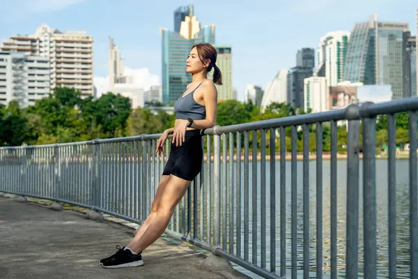 Asyalı bir kadın duvarlara yaslanmış, arka planda büyük bir şehir ve yumuşak bir ışıkla halka açık alanda egzersiz yaptıktan sonra rahatlamak için saatine bakın..
