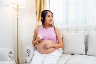 Asyalı hamile kadın kanepeye otur ve bir bardak süt tut ve karnına dokun ayrıca mutlulukla sağ tarafına bak..