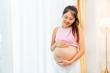 Asyalı hamile bir kadın, beyaz perdenin önünde karnına dokunup mutlu bir şekilde gülümsüyor..