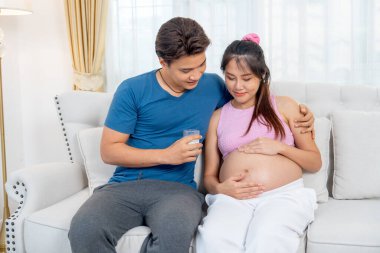 Asyalı bir adam hamile bir kadının yanında kanepede oturur ve elinde bir bardak süt ile kadına sarılır ve sevgisini ifade eder..