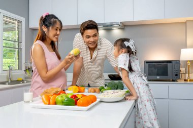 Asyalı hamile anne, yanında duran küçük kız ve babaya mısır gösterip oyun oynuyor. Mutfakta birlikte mutlu görünüyorlar..