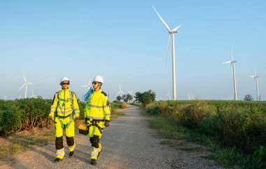 Rüzgâr türbini ya da yel değirmeni işçisi ya da teknisyenlerinden ikisinin geniş açılı görüntüsü araçlar taşıyor ve iş yerinde arka planda bir sıra rüzgar türbiniyle yol boyunca yürüyor.