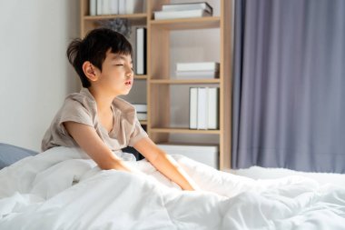 Asyalı çocuk sabah uyanır uyanmaz yatağa oturur ve gözlerini sabah ışığıyla uykuya daldırır..