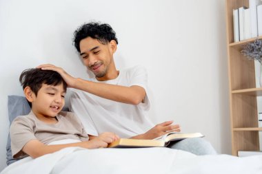 Asyalı genç bir baba yatakta birlikte yatarken oğlunun kafasına dokunur ve mutlu bir şekilde kitabı okur..