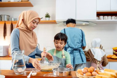 Muslim mother stand beside of her son to mix flower in glass bowl and the other also cook in the back of kitchen in their house. clipart