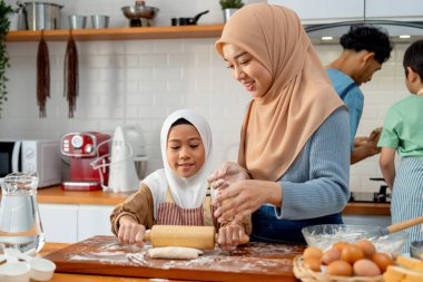 Little Asian muslim girl enjoy to use rolling pin to make some bakery and support beside by her young mother in the kitchen with father and brother also cooking in the back. clipart