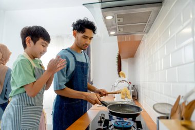 Side view young Asian father show skill of cooking by throwing pan cake up and his son cheer up beside in the kitchen of their house. clipart