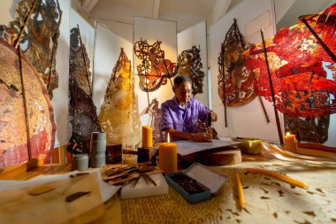 Asyalı kıdemli bir adamın yan görünüşü, çekiç ve keski kullanarak büyük gölgeler yaratması ve işyeri çevresinde çeşitli ürünlerin bulunduğu bir alanda çalışmaya odaklanması.