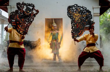 Khon maskeli bir adam melek olarak eski binanın kapısının önünde duruyor. İki adam da büyük gölgeyi tutuyor. Önde oynuyorlar ve arkada sıcak bir ışık var..