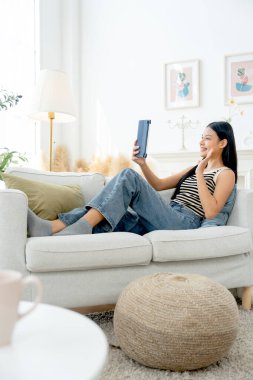 Güzel Asyalı kadının dikey görüntüsü kanepeye yaslanarak oturma odasında tablet kullanmaktan zevk alır ve gün ışığında heyecanını ve mutluluğunu ifade eder..
