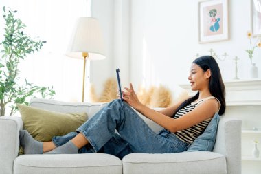 Yan görüş ve geniş açı. Güzel Asyalı kadın kanepeye yaslanıyor. Oturma odasında gün ışığında tablet kullanmaktan da zevk alıyor..