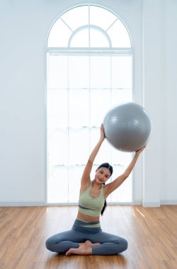 Egzersiz elbiseli güzel bir Asyalı kadının dikey görüntüsü otur ve yoga egzersizi yap. Gri topu tut ve sol tarafına kaldır ve cam pencerenin önünde kal..