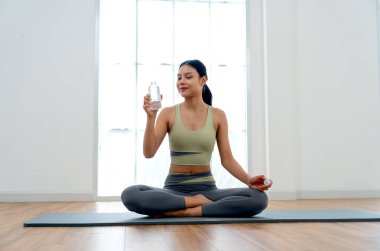 Egzersiz elbiseli güzel bir Asyalı kadın su şişesini tutuyor ve yoga minderine oturuyor ve cam pencereden gün ışığına çıkıyor..
