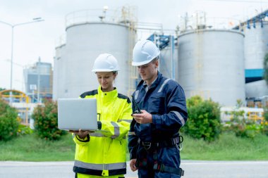 Petrokimyalı erkek ve kadın işçiler veya mühendisler fabrika tankının önünde dizüstü bilgisayarlarını kullanarak işlerini tartışıyorlar..