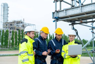 Bir grup petrokimya işçisi ya da teknisyen bir dizüstü bilgisayarla birlikte çalışıyorlar ve fabrikanın önünde duruyorlar ve işyerinde mutluluğa bakıyorlar..