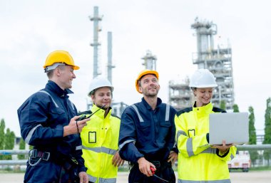 Dört petrokimya işçisi veya teknisyen bir dizüstü bilgisayarla birlikte çalışıyor ve fabrikanın önünde duruyorlar..