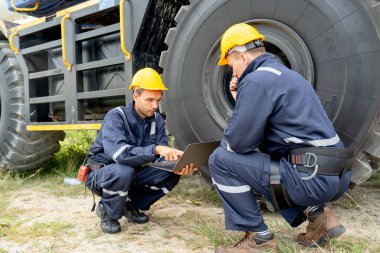 Bir fabrika işçisi dizüstü bilgisayar kullanıyor ve kamyonun direksiyonuna yakın oturuyor ve iş yerindeki iş arkadaşıyla kontrol ya da bakım yapıyor..