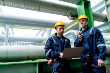 Petrokimya işçisi telsizi elinde tutuyor ve iş arkadaşıyla bilgisayarını tutuyor ve fabrika işyerindeki boru hattında kalıyor..