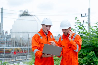 Bir mühendis ya da gaz fabrikası işçisi dizüstü bilgisayarı işaret edip telsiz kullanan diğeriyle konuşur ve iş yerindeki benzin deposunun önünde dururlar..