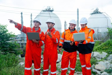 Bir dizüstü bilgisayarı olan bir grup gaz fabrikası teknisyeni veya mühendisi ve bir tanesi sağ tarafını göstererek çalışmalarını tartıştılar ve gün ışığında tankın önünde durdular..