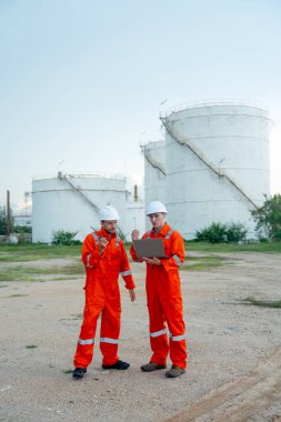 İki mühendis veya gaz fabrikası işçisinin iş yerindeki gaz veya petrokimya deposu önünde dizüstü bilgisayarla tartışmalarının dikey görüntüsü.