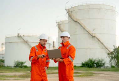 İki mühendis veya gaz fabrikası çalışanı, iş yerindeki gaz veya petrokimya deposu önünde dizüstü bilgisayarı kullanarak tartışırlar..