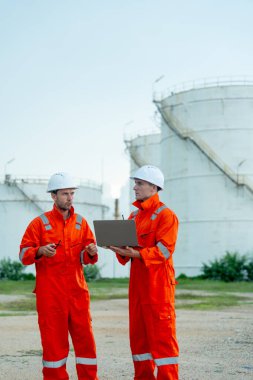 İki mühendisin ya da gaz fabrikası teknisyeninin dikey görüntüsü ellerinde telsiz ve dizüstü bilgisayar ile yaptıkları işleri tartışıyor ve tankın önünde duruyorlar..