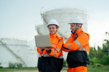 Teknisyen ya da gaz fabrikası işçisi olan kadın, gaz ya da petrokimya deposu önünde iş arkadaşıyla çalışmak için dizüstü bilgisayarı tutuyor ve kullanıyor ve işlerinde mutlu görünüyorlar..