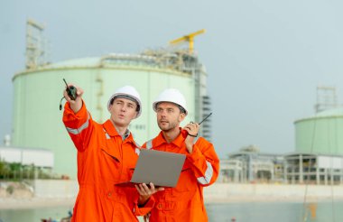 Bir gaz fabrikası çalışanı sağ tarafını telsizle gösteriyor ve sorunu çözmek için iş arkadaşıyla konuşuyor ve körfezin yanındaki benzin deposunun önünde duruyor..