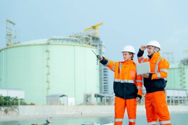 Petrokimya veya gaz fabrikası işçileri veya teknisyenleri fabrikanın önünde çalışıyor. Bir kadın sağ tarafını gösteriyor, bir erkek dizüstü bilgisayarı tutuyor ve kadınlara bakıyor..