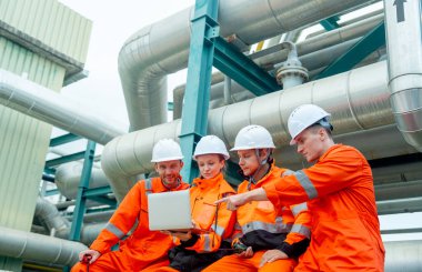 Bir grup petrokimya ya ya da gaz fabrikası işçisi ya da mühendis laptopu kullanarak iş hakkında tartışıyor ve fabrika işyerindeki boru hattının yanında oturuyor.