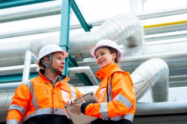 İki petrokimyasal ya da gaz fabrikası işçisi kadın ve erkek bilgisayar kullanarak iş hakkında konuşuyor ve fabrika işyerindeki boru hattının yanında duruyorlar..