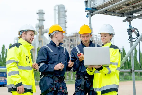 Bir grup petrokimya işçisi ya da teknisyen bir dizüstü bilgisayarla birlikte çalışıyor ve fabrikanın önünde duruyorlar..