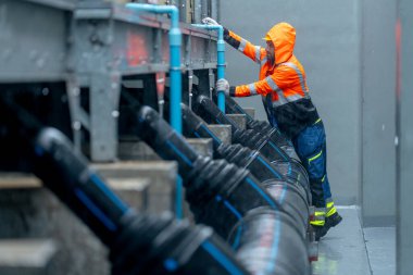 Yağmurluklu fabrika işçisi veya teknisyeninin yan görüntüsü boru sistemi boyunca yürüyor ve yağmur yağarken fabrika binasının bakımına bakıyor.