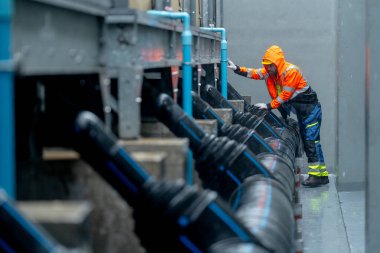 Yağmurluklu fabrika işçisi ya da teknisyen geniş açıda boru sistemi boyunca yürüyor ve yağmur yağarken fabrika binasının bakımını üstleniyor..