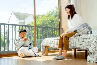 Asyalı anne yatağa oturur ve elma yemekten hoşlanan oğluna bakar. Sabah ışığı ve mutlulukla yatak odasında eskrim sporu yaparken de yere oturur..