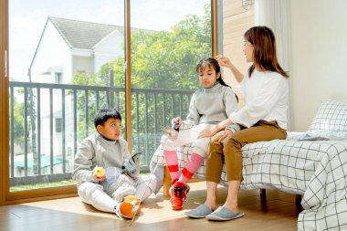 Asyalı anne, kızının yanına, kızının ve oğlunun saçlarına dokunup sabah ışığı ve mutlulukla yatak odasında eskrim sporu yaparken yere oturur..