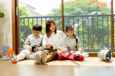 Asyalı anne, oğul ve kız aile olarak cam kapının önüne oturup kızla rahatlayın ve oğlanın cep telefonunun keyfini çıkarın..