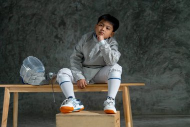 Eskrim üniformalı Asyalı çocuk çişini ve maskesini yanına koydu ve antrenmandan sonraki rahatlama sırasında ahşap bankta oturup yarışmayı düşündü..