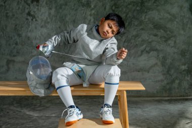 Eskrim üniformalı Asyalı çocuk çişini tut ve bük ayrıca ahşap bankta otur ve antrenmandan sonraki rahatlama sırasında yarışmayı düşünüyor gibi görün..