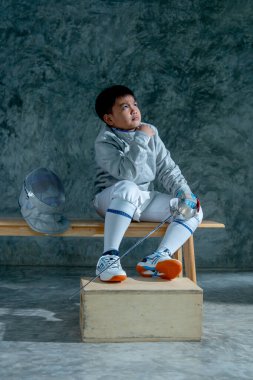 Çocuğun dikey görüntüsü, eskrim üniforması giyer, çişini tutar ve tahta bir bankta oturup sol tarafına bakar..