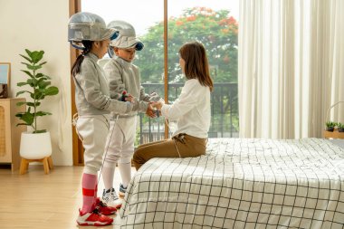 Asyalı anne yatakta oturur ve çocuğunun üniformasını ve aletlerini evde eskrim sporu yapmak için alır..