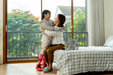 Asyalı anne yatakta kızıyla birlikte oturur ve eskrim antrenmanı sırasında yumuşak sabah ışığı ve mutluluğu ile yatak odasında birbirlerine sarılırlar..