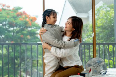 Asyalı anne yatak odasında sabah ışığı ve mutlulukla eskrim sporu yaparken kızıyla birlikte yatağa oturup sarılıyor..