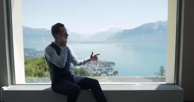 Genç işadamı cep telefonuyla konuşuyor. Pencerenin yanında, göl ve dağların panoramik manzaralı. 
