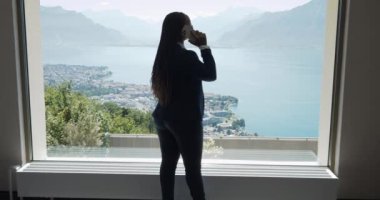 Genç Afrikalı iş kadını, pencerenin yanındaki cep telefonuyla konuşuyor. Gölün ve dağların panoramik manzarası var. 