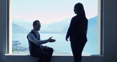 Genç ve başarılı iş adamları toplantı yapıyor. Erkek ve kadın pencerenin yanındaki modern ofiste nefes kesici bir manzarayla ayakta duruyor ve konuşuyor.