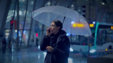 Kızgın genç kadın, yağmurlu bir şehirde gece vakti telefonda konuşuyor.
