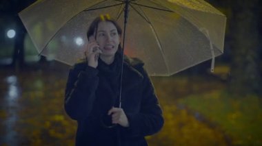 Kızgın genç kadın, yağmurlu bir şehirde gece vakti telefonda konuşuyor.